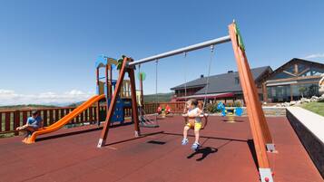 Un parque infantil con columpios y toboganes, una estructura de madera, y un edificio al fondo.