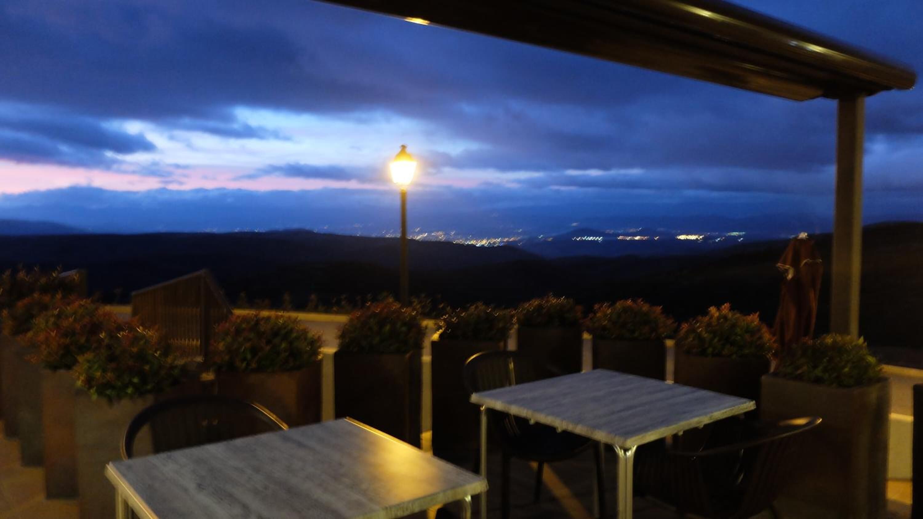 Una terrassa a l'exterior amb taules i cadires, un fanal i vista a un paisatge urbà a la capvespre.
