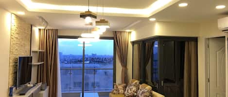 Una sala moderna con un sofá, una mesa de comedor y un televisor. Hay un ventanal con cortinas, y la habitación tiene vista a la ciudad.
