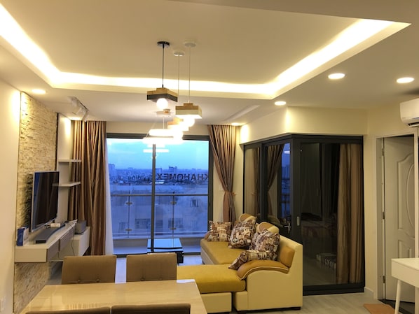 A modern living room with a sofa, a dining table, and a TV. There is a large window with curtains, and the room has a city view.