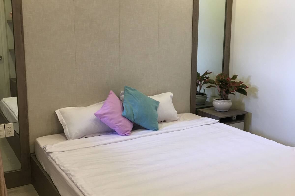 Un lit bien fait, avec du linge de lit blanc et des coussins colorés, une table de chevet ornée d’une plante en pot et un grand miroir accroché au mur.