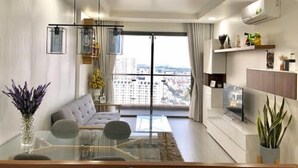 A modern living room with a sofa, a glass coffee table, and a dining area with a glass table. There is a large window with a view of buildings outside. - The GoldView Luxury Apartment (Ho Chi Minh City)