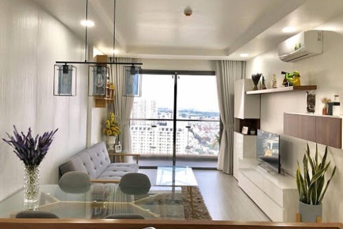 Un salon moderne avec un canapé, une table basse en verre et un coin repas doté d’une table en verre. Une grande fenêtre offre une vue sur les bâtiments environnants.