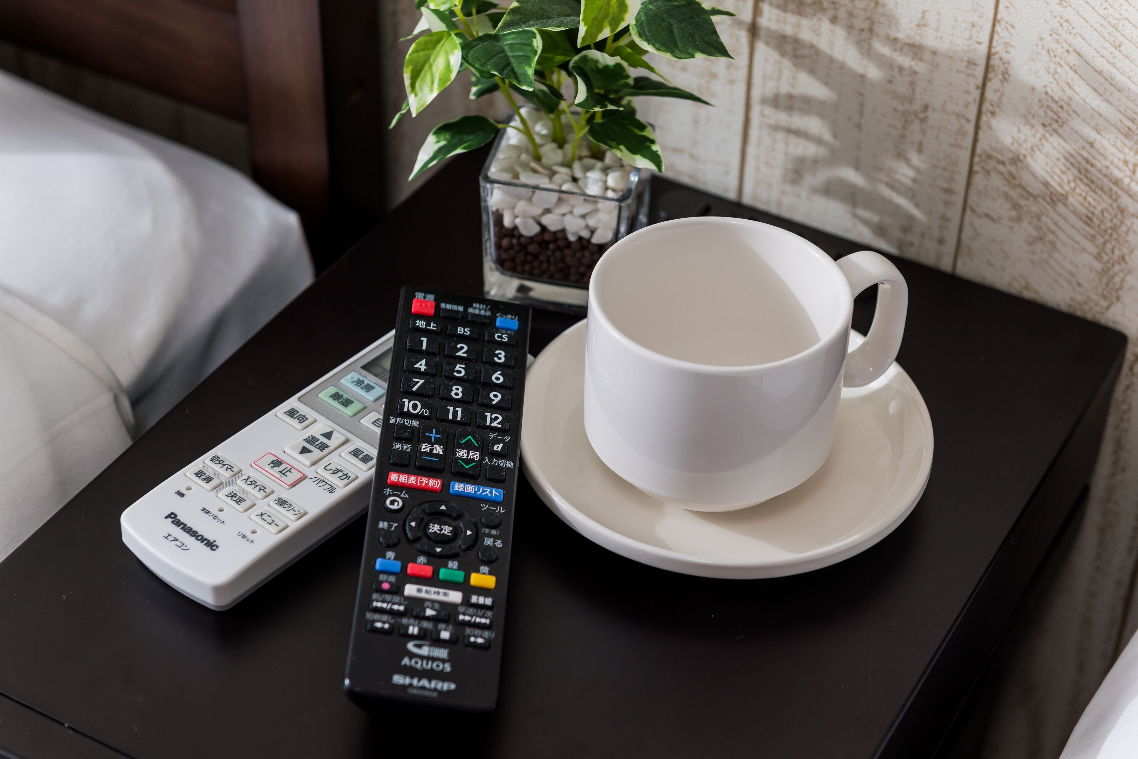 Un control remoto, una taza blanca y una planta pequeña en una mesa.