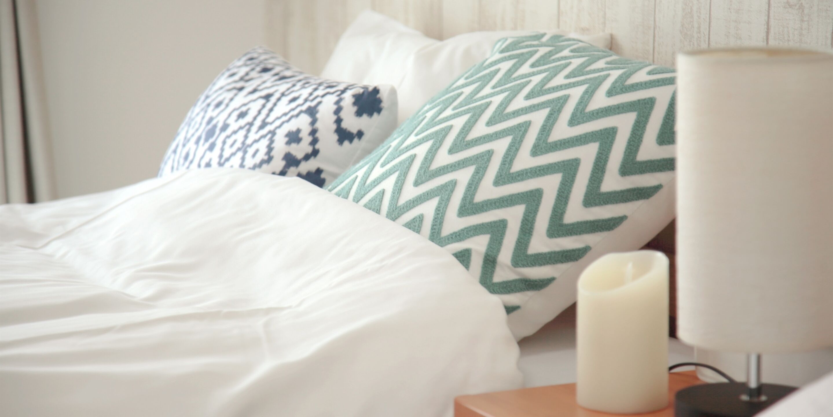 Un lit bien fait, avec une literie blanche et des coussins décoratifs, une table de chevet sur laquelle se trouve une bougie blanche et une lampe.