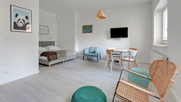 Quarto moderno e bem iluminado, com cama, mesa de jantar com cadeiras, TV fixada na parede e uma cadeira de vime.