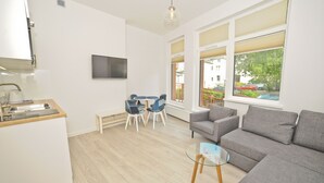 Uma cozinha moderna com forno embutido, um espaço de refeições com mesa e cadeiras, e um sofá cinzento numa sala de estar luminosa com grandes janelas.