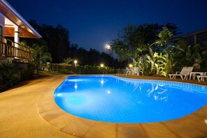 Ein Schwimmbad im Abenddämmerung mit Mond im Hintergrund, umgeben von Palmen und Liegestühlen.