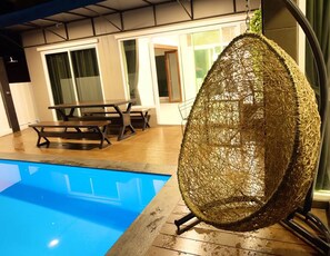 A hanging chair near a pool with a wooden deck and a table with chairs in the background. - Moon Terrace Pool Villa (Hua Hin)