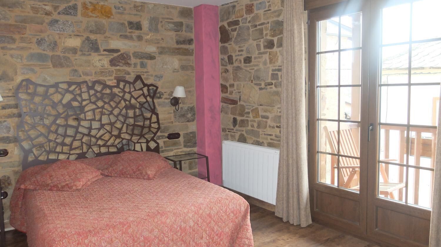 A bedroom with a stone wall, a bed with a pink bedspread, a window with a view of outdoor furniture, and a lamp on the wall.