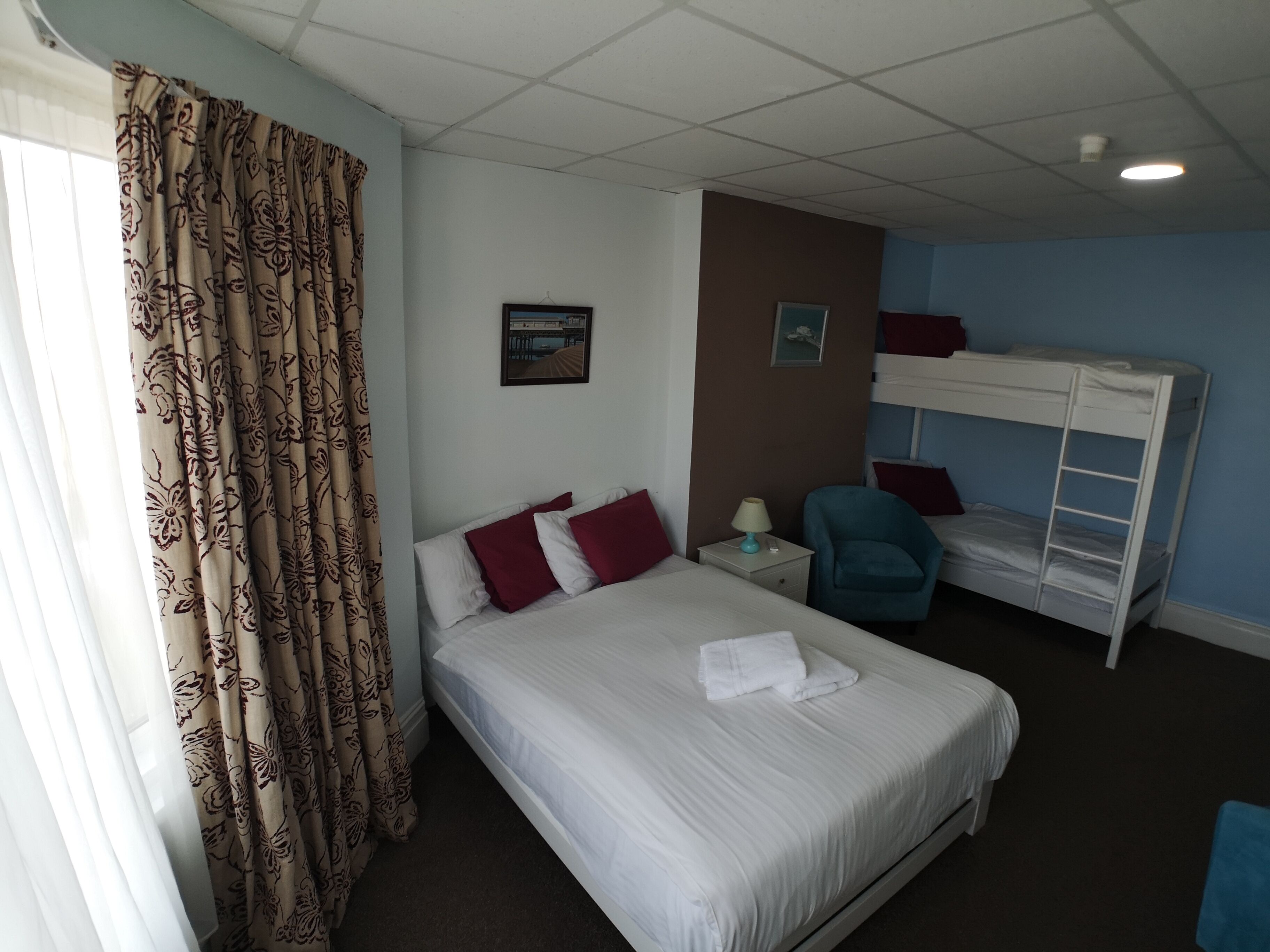 A hotel room with a large bed, a bunk bed, a chair, a nightstand, and a window with curtains.