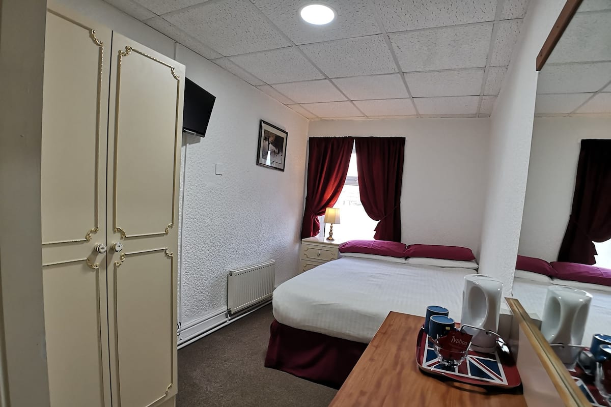 A hotel room with a bed, a wooden desk, a dresser, a mirror, a radiator, and a TV on the wall.