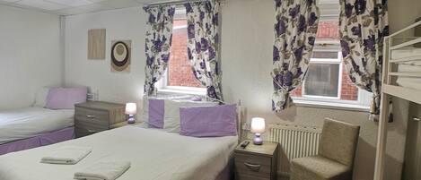 A hotel room with two beds, a chair, a nightstand with a lamp, and a radiator.