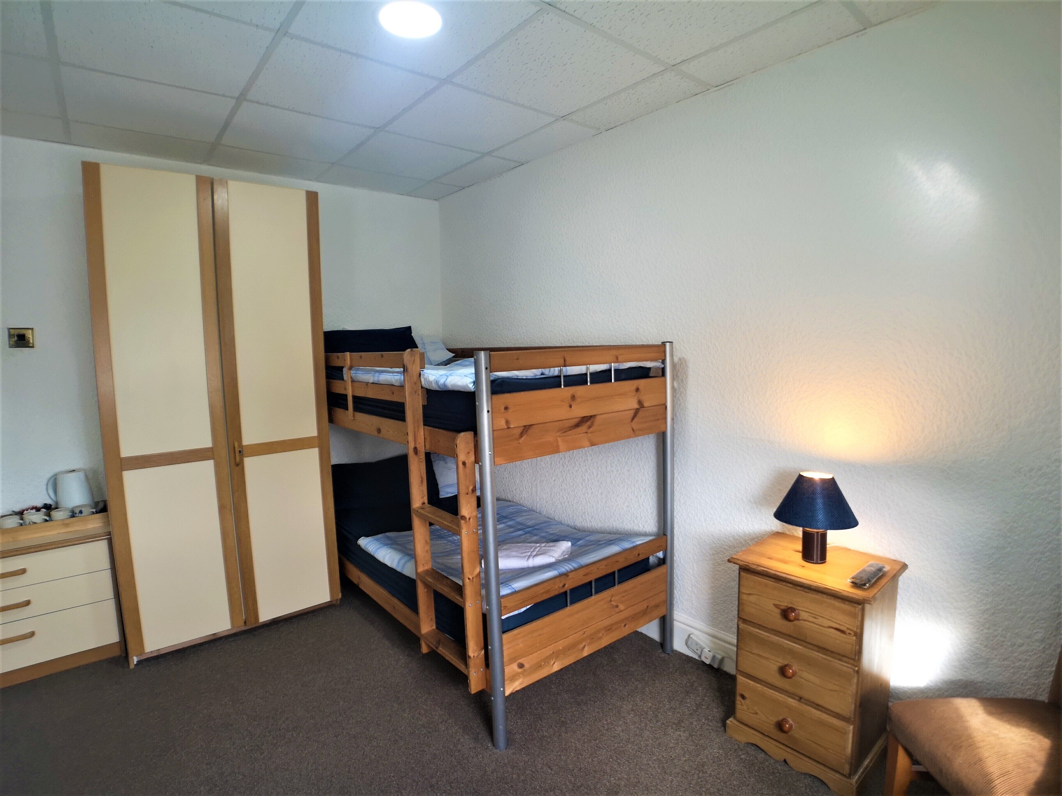 Un lit superposé avec un bureau et une commode, une lampe et une chaise, dans une pièce aux murs blancs et dotée d’un éclairage au plafond.