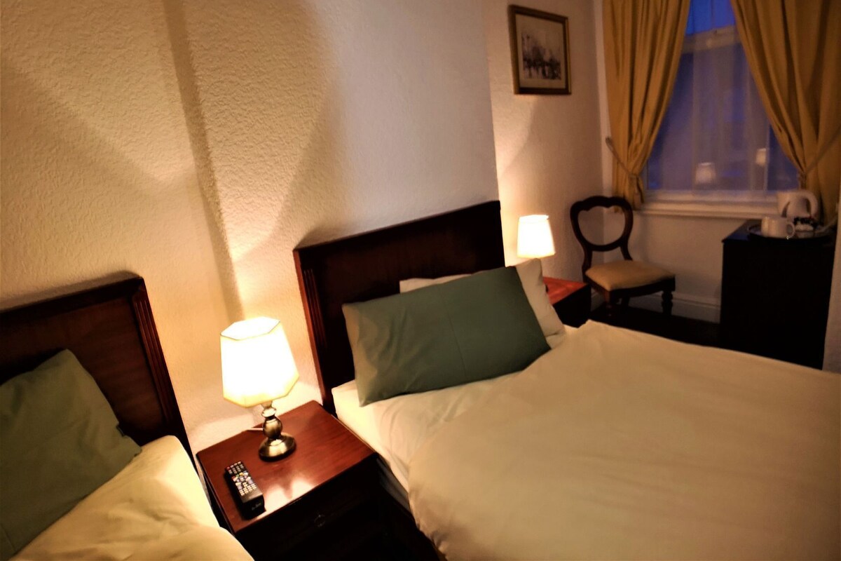A hotel room with two beds, a nightstand with a lamp, a chair, a desk with a coffee pot, and a window with curtains.