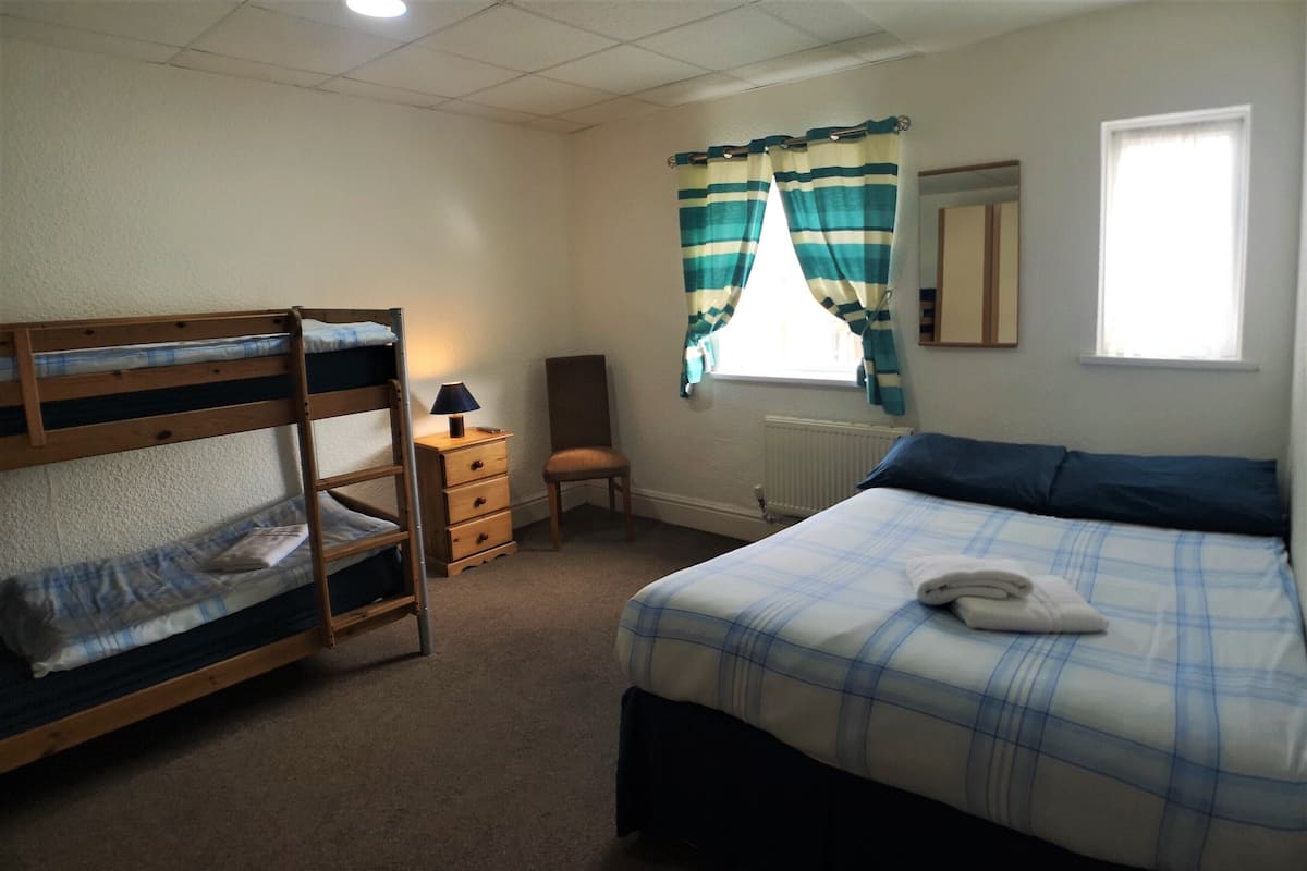 A dormitory room with bunk beds, a single bed, a chair, a nightstand, a lamp, a window with curtains, and a radiator.