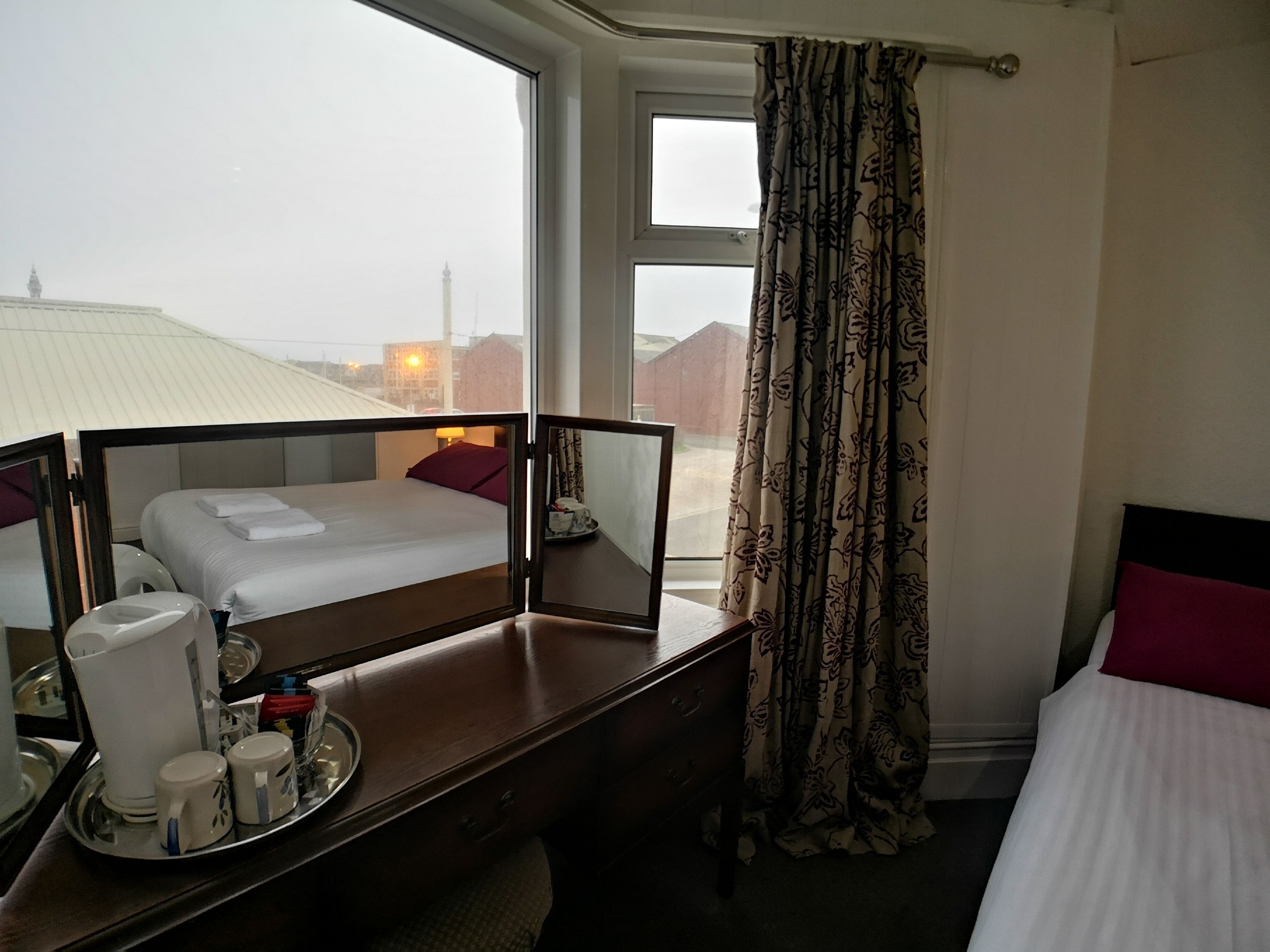 Habitación de hotel con cama, escritorio con una bandeja de objetos, ventana con cortinas y vista de edificios.