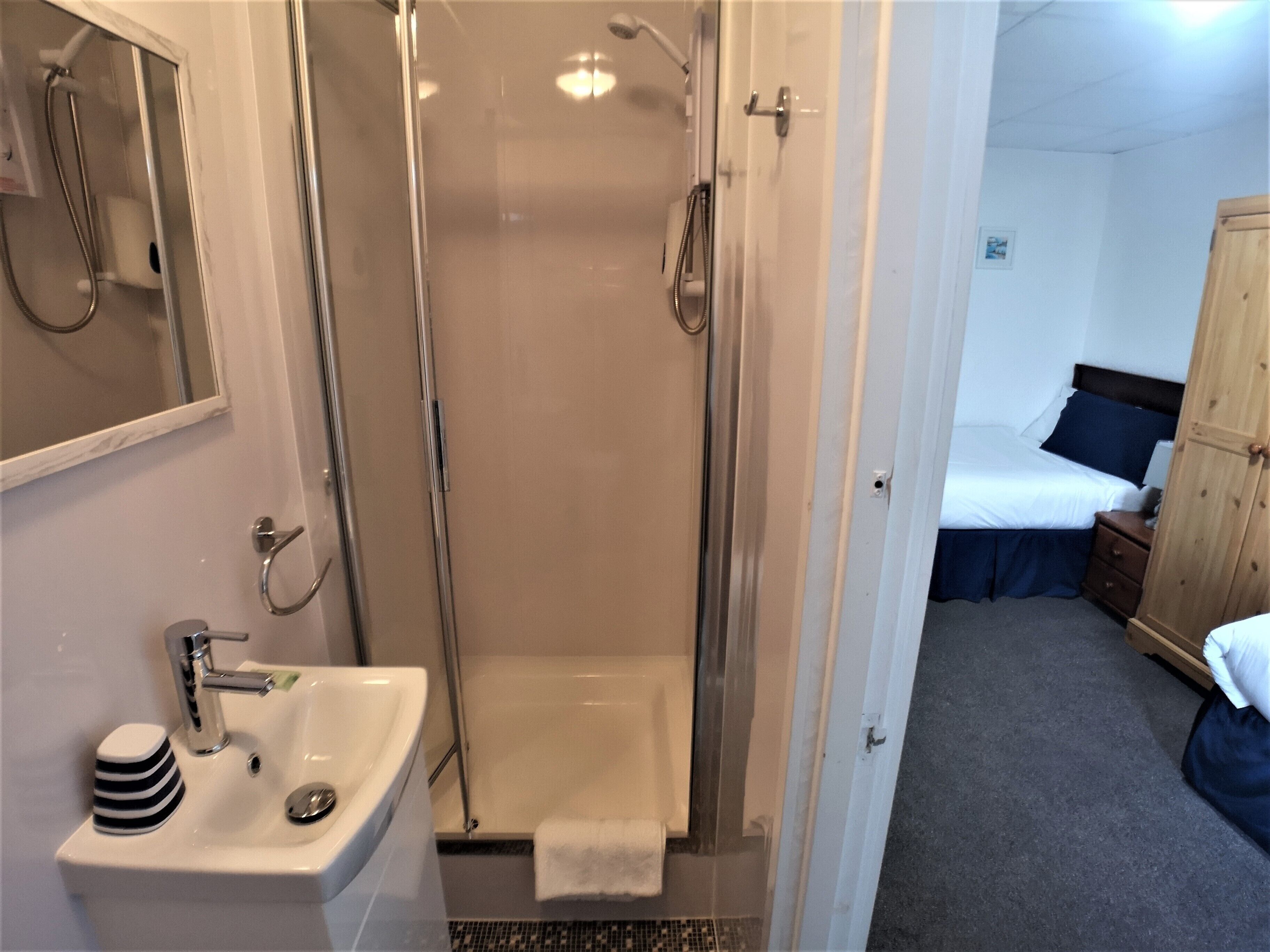 Baño con ducha, lavabo y espejo. En la habitación contigua, hay una cama con un almohadón azul.