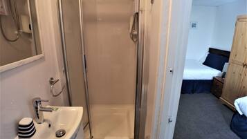 Baño con ducha, lavabo y espejo. En la habitación contigua, hay una cama con un almohadón azul.
