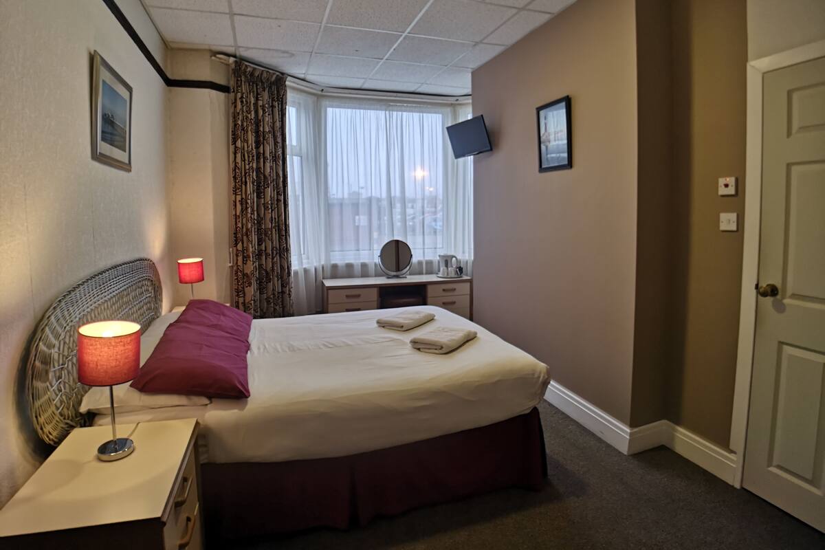 A hotel room with a bed, bedside lamp, dresser, vanity, and a window with curtains.