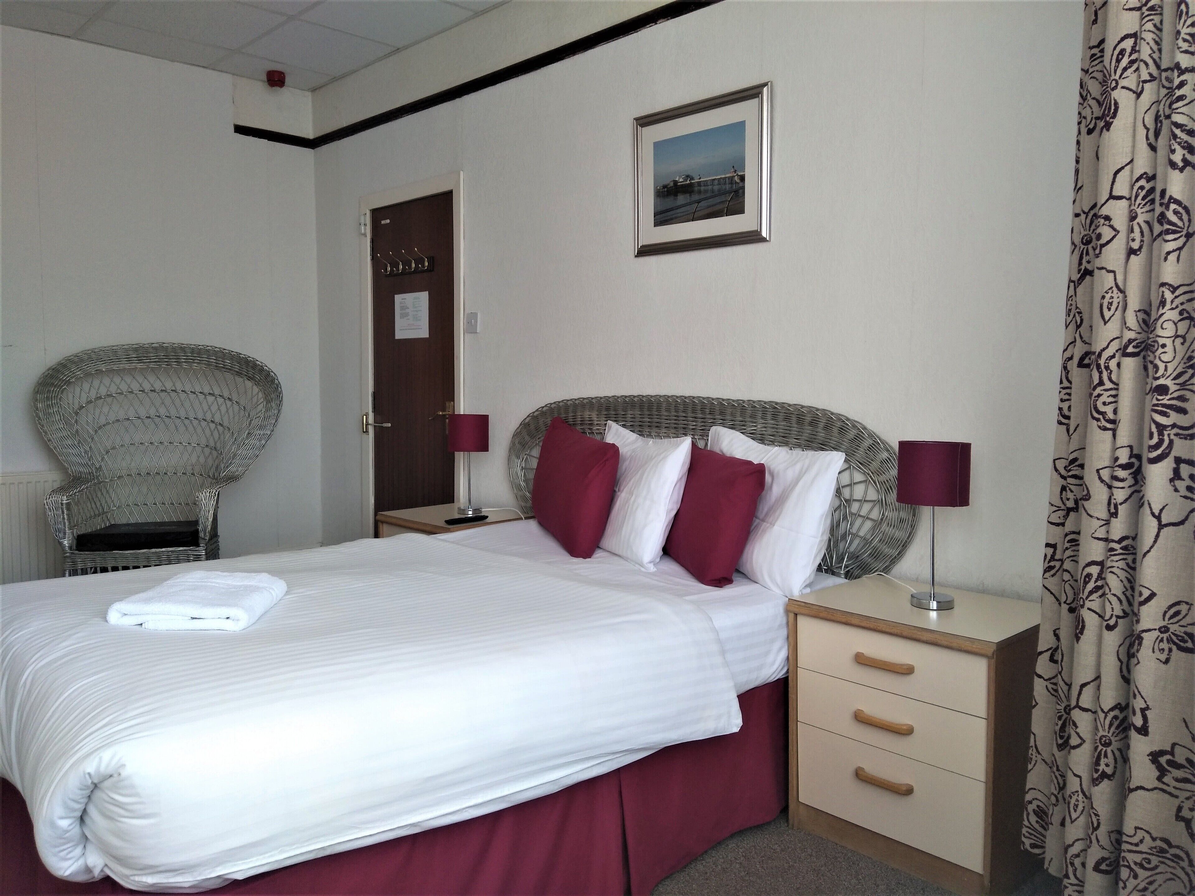 A hotel room with a bed, a wicker chair, a nightstand with a lamp, and a door with a sign.