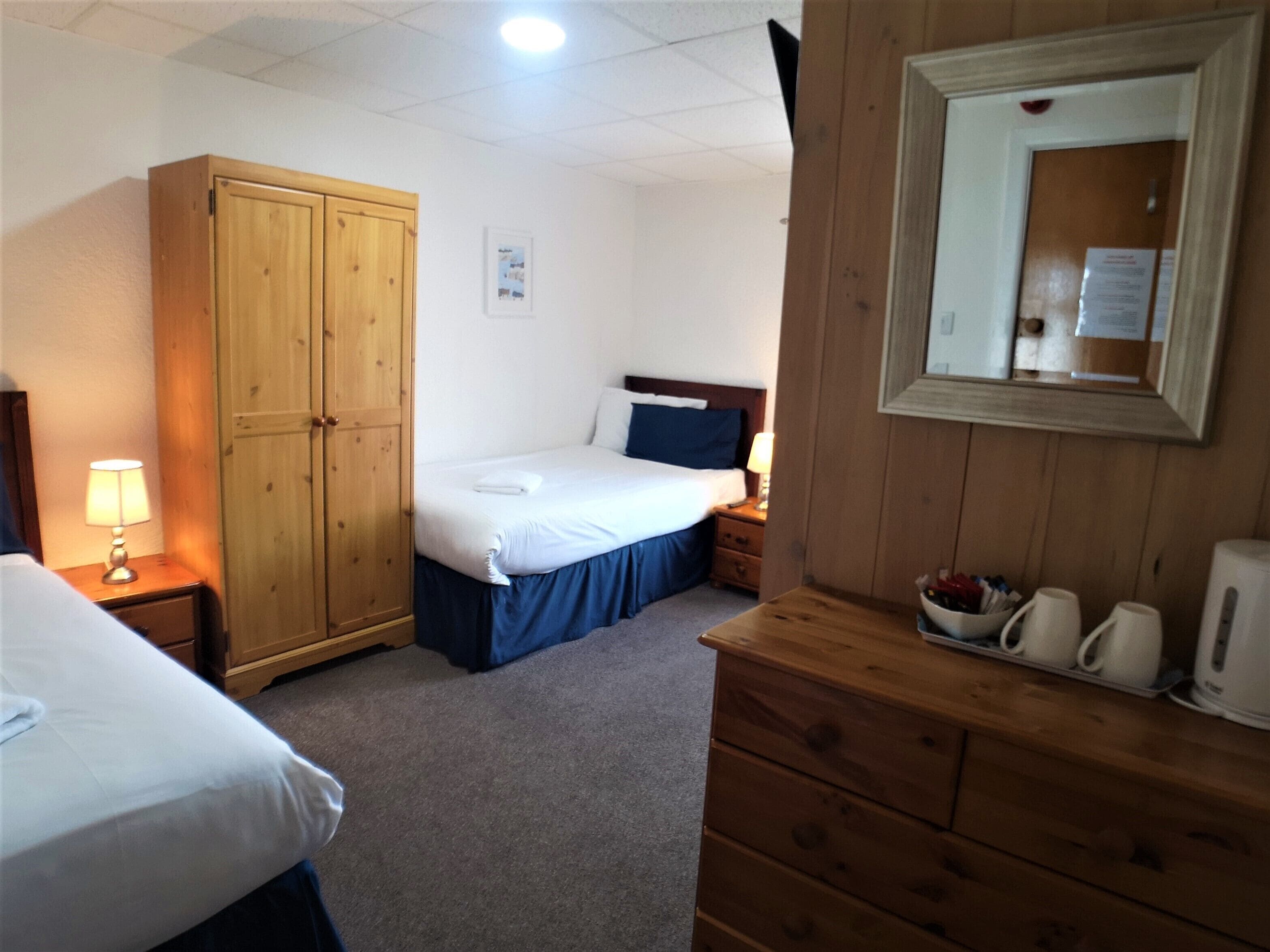 A hotel room with a bed, a wardrobe, a nightstand with a lamp, and a dresser with cups and a kettle.