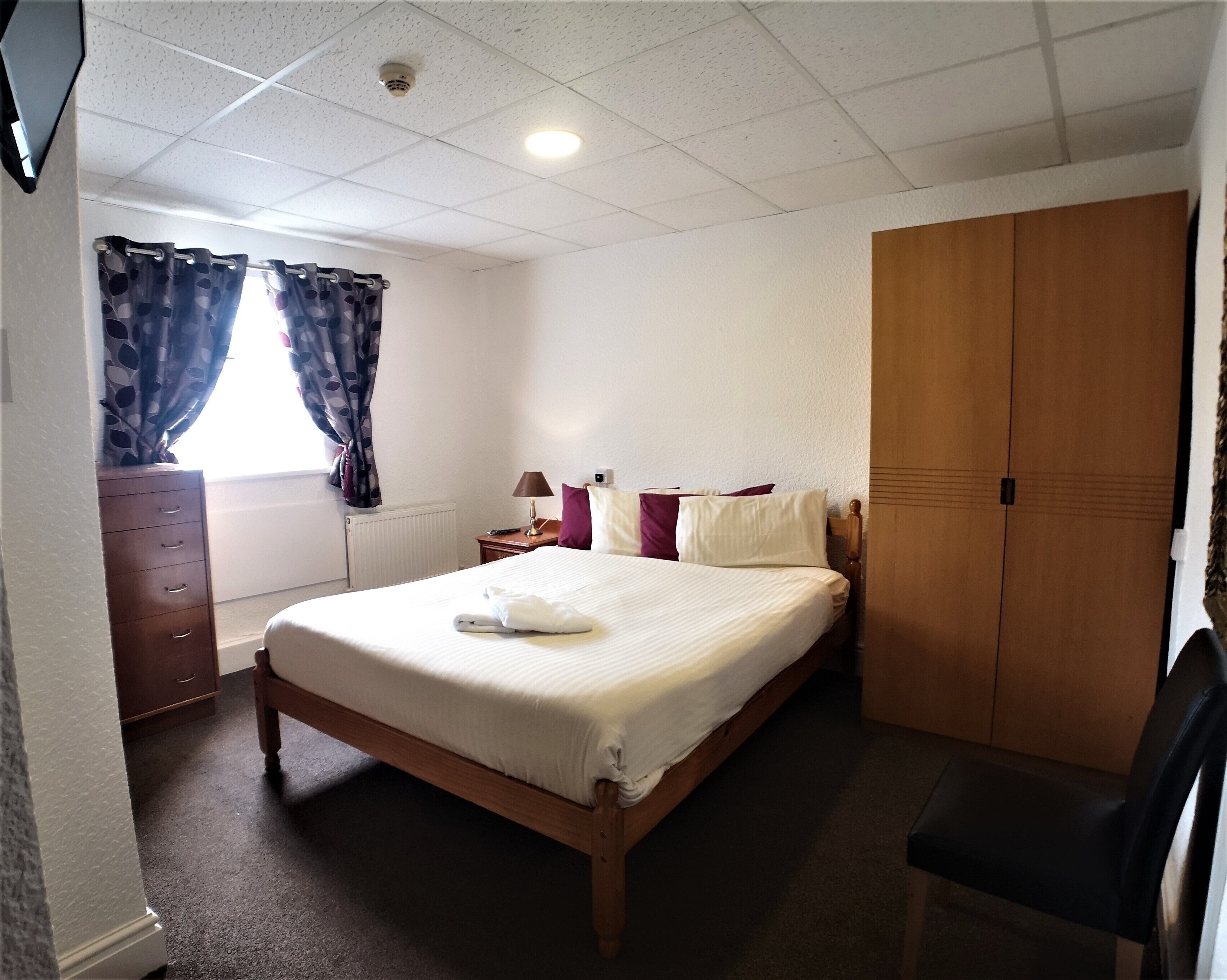 A hotel room with a bed, a wooden wardrobe, a desk with a lamp, and a chair.
