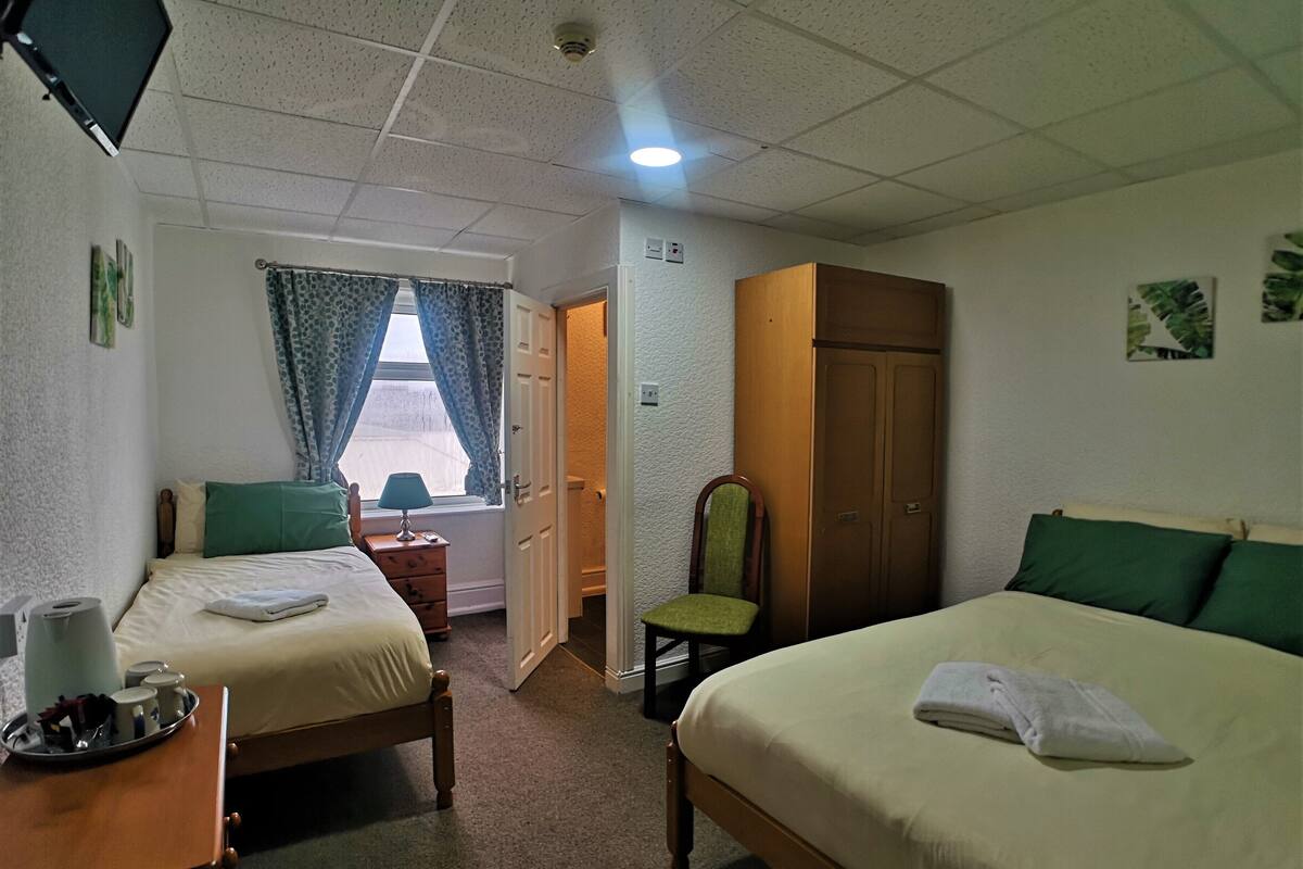A hotel room with two beds, a wooden wardrobe, a desk with a lamp, and a window with curtains.
