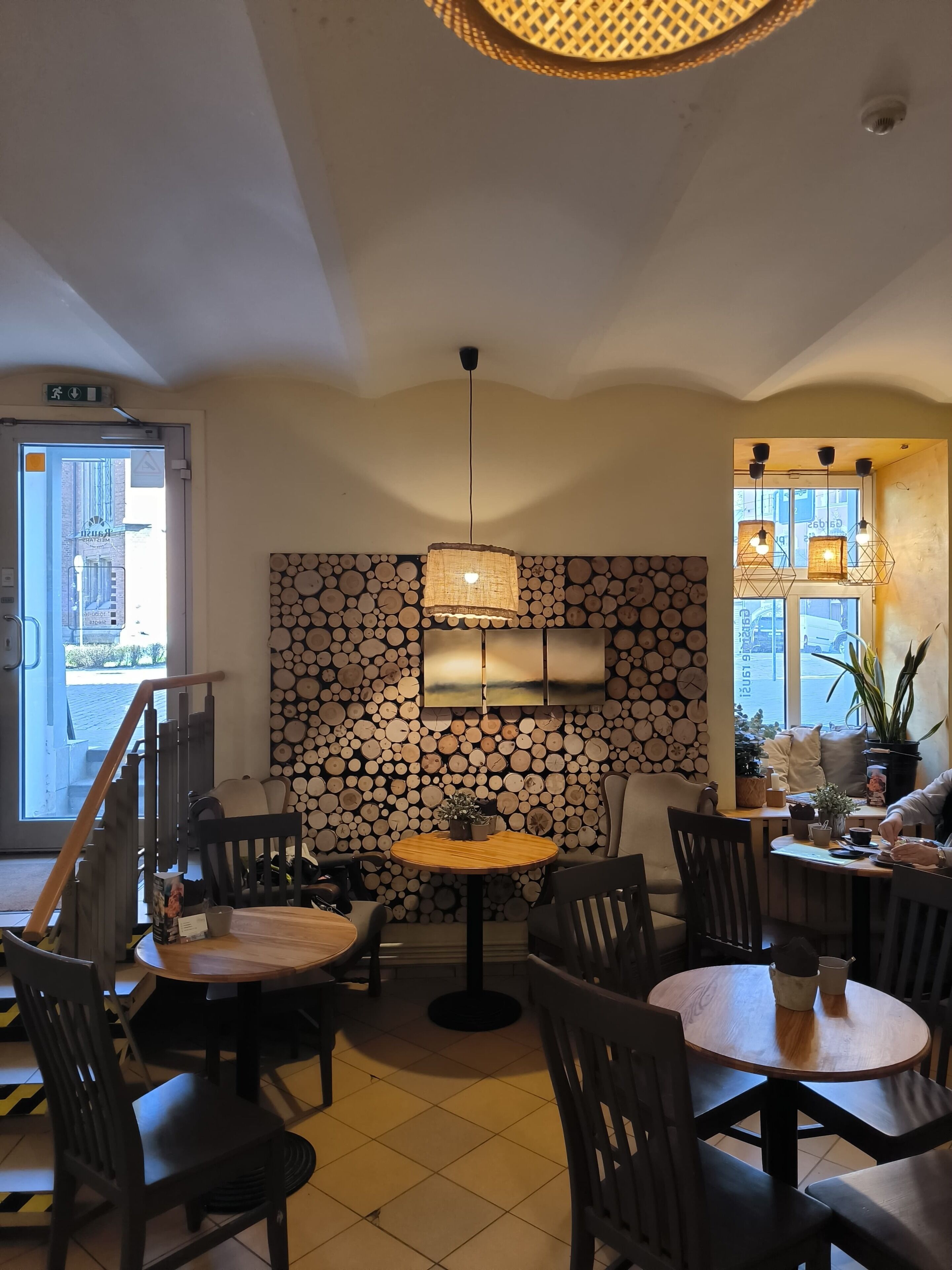 Un acogedor interior de cafetería con mesas redondas, sillas, una pared de troncos de madera y lámparas colgantes.