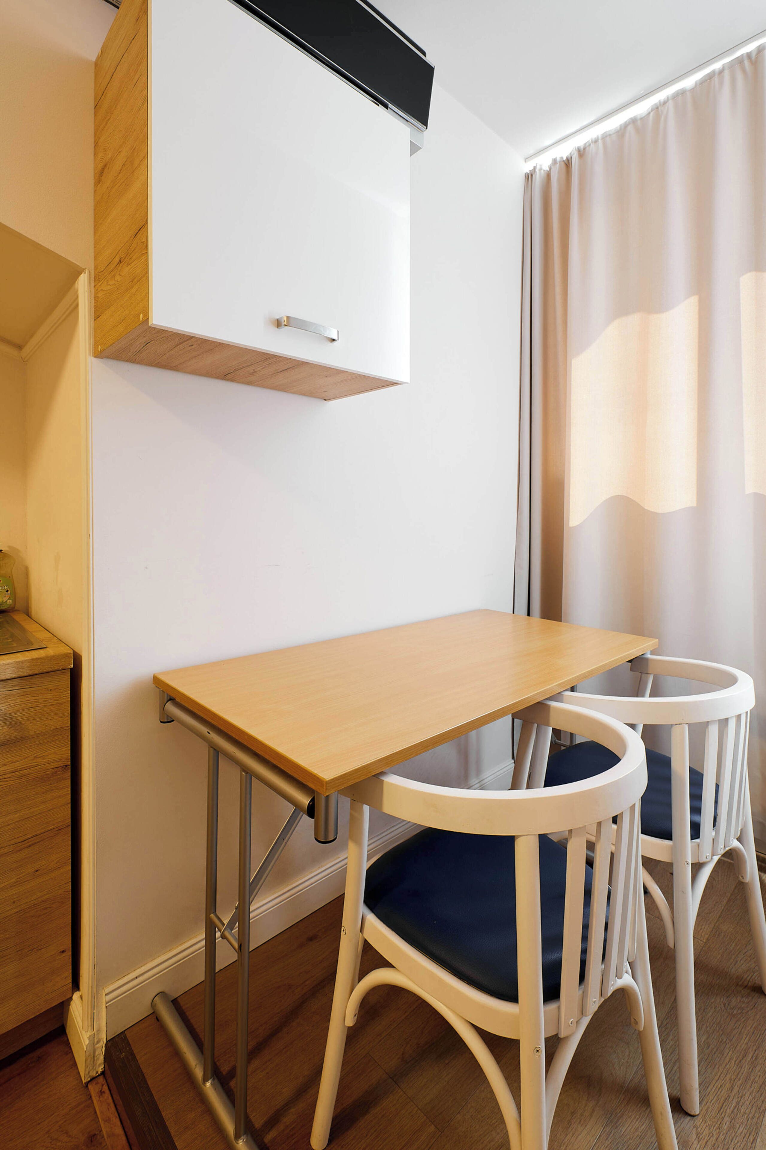Une petite pièce avec une table en bois, deux chaises blanches dotées de coussins bleus, une armoire blanche et une fenêtre avec des rideaux.