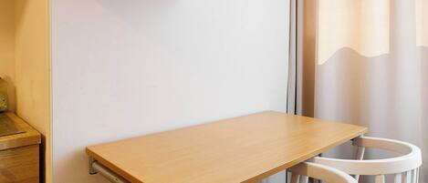 Une petite pièce avec une table en bois, deux chaises blanches dotées de coussins bleus, une armoire blanche et une fenêtre avec des rideaux.