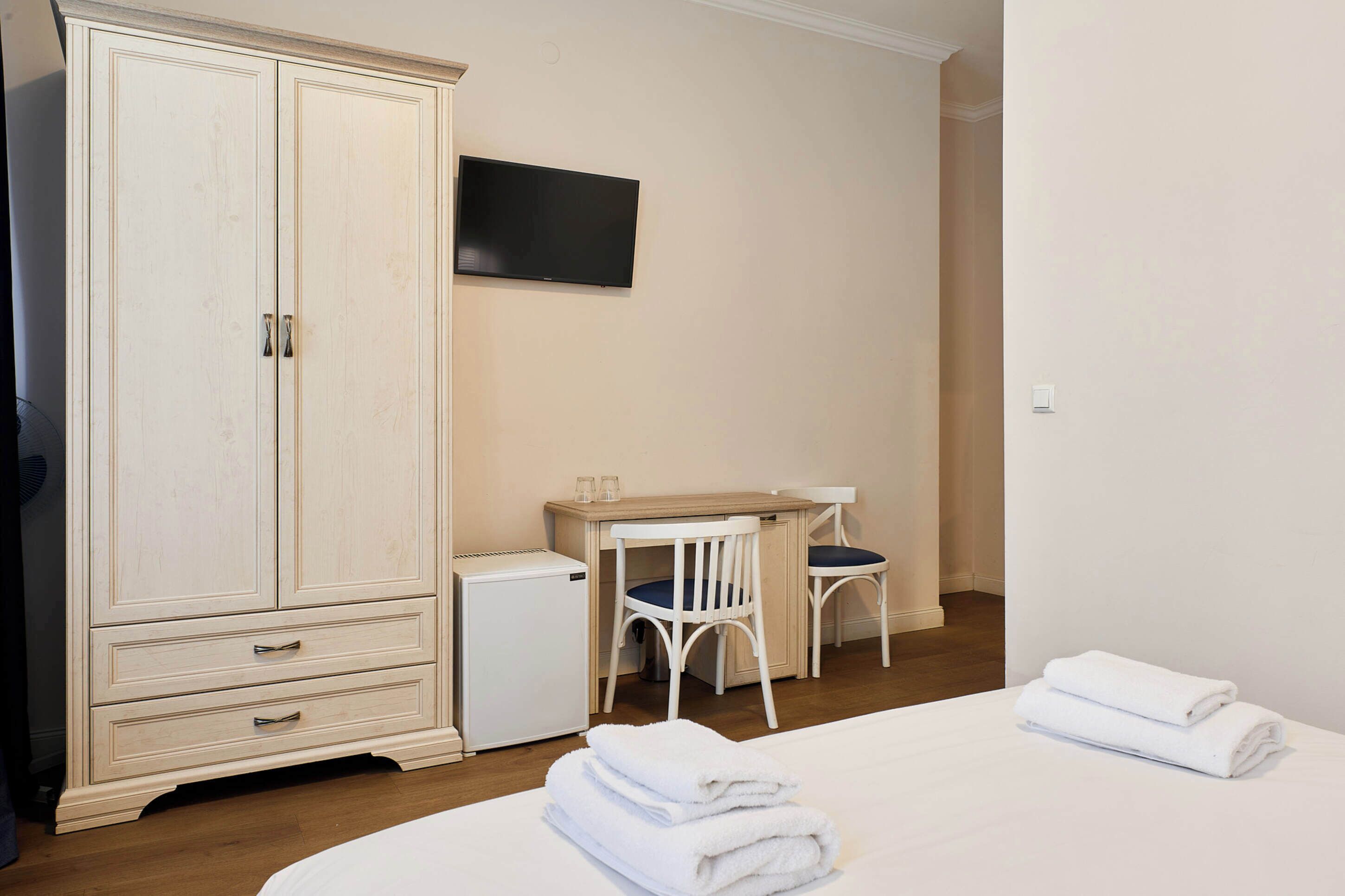 Habitación de hotel con un armario grande, un escritorio pequeño con dos sillas, un televisor en la pared y toallas ordenadamente dobladas sobre la cama.