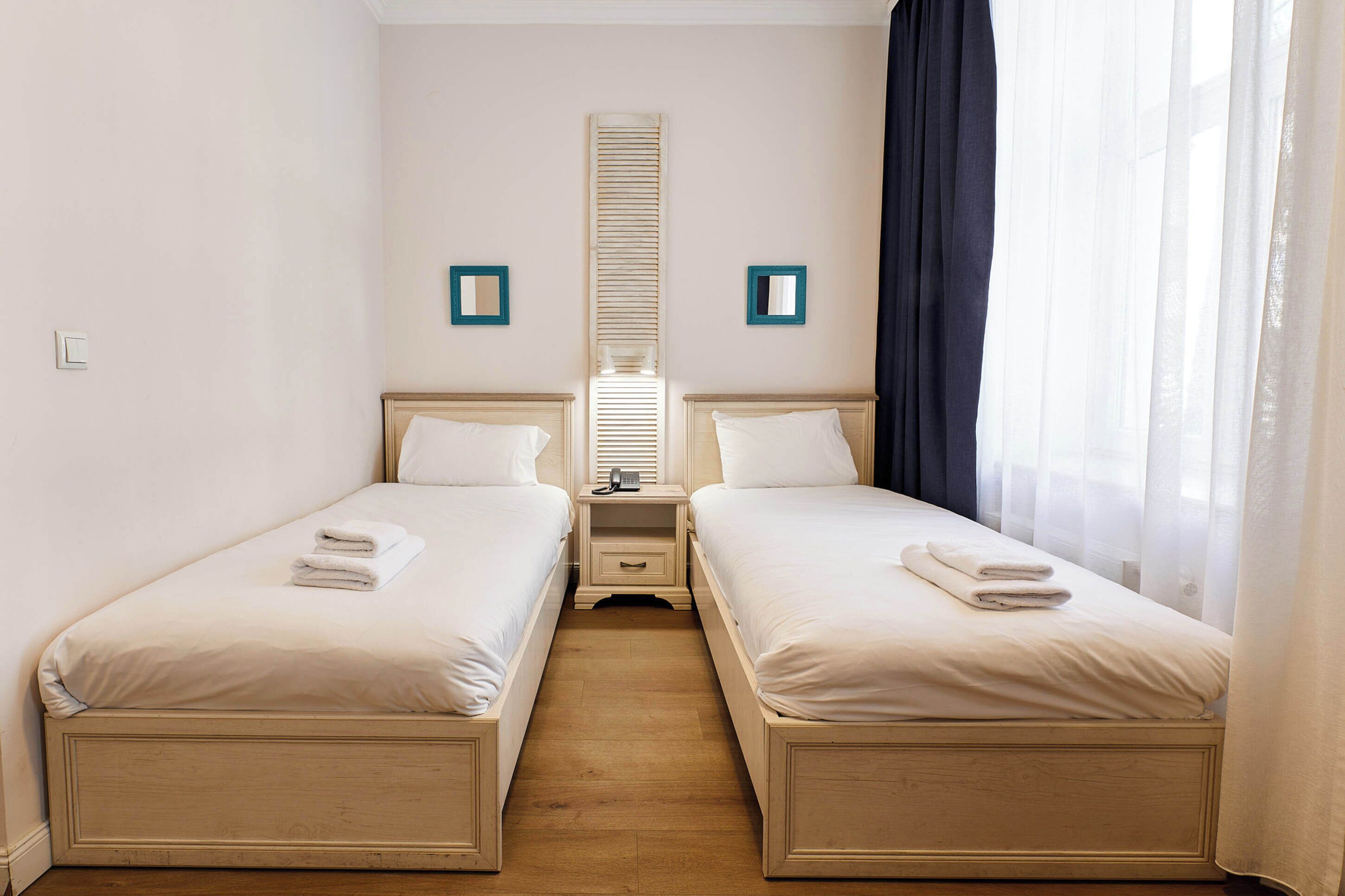 Ein Doppelzimmer mit zwei Betten, einem kleinen Nachttisch mit Lampe, einem Spiegel und einem Fenster mit transparenten Vorhängen.