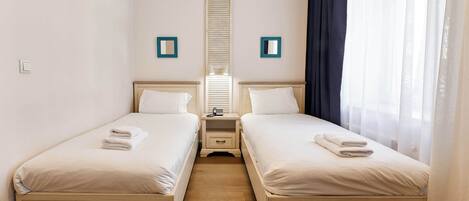 A twin room with two beds, a small bedside table with a lamp, a mirror, and a window with sheer curtains.