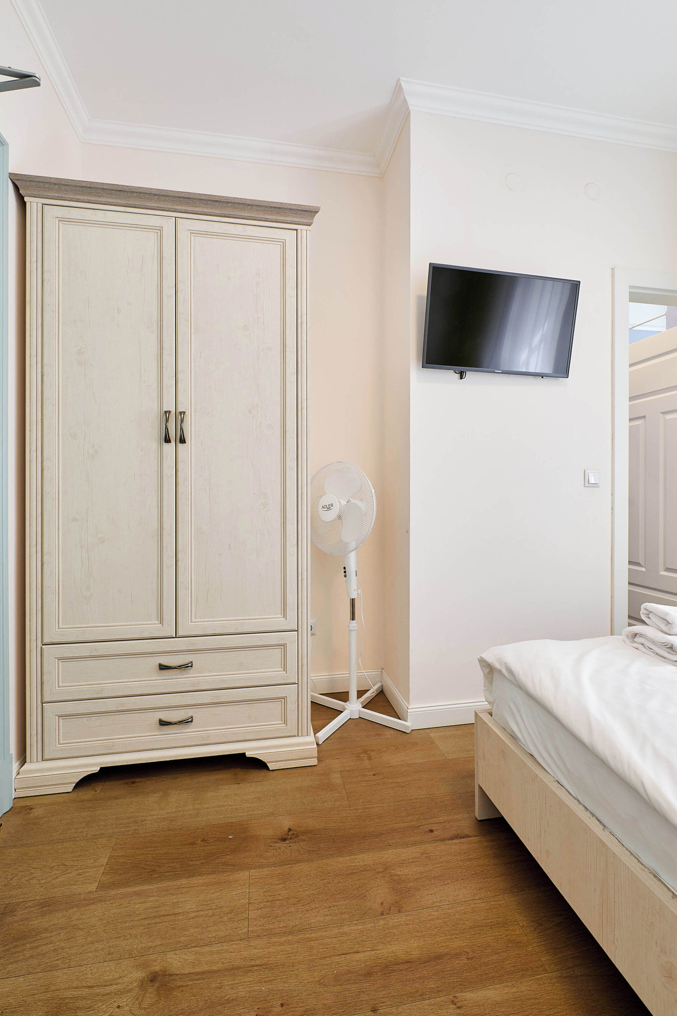 A bedroom with a wooden floor, a bed, a wardrobe, a wall-mounted TV, and a standing fan.