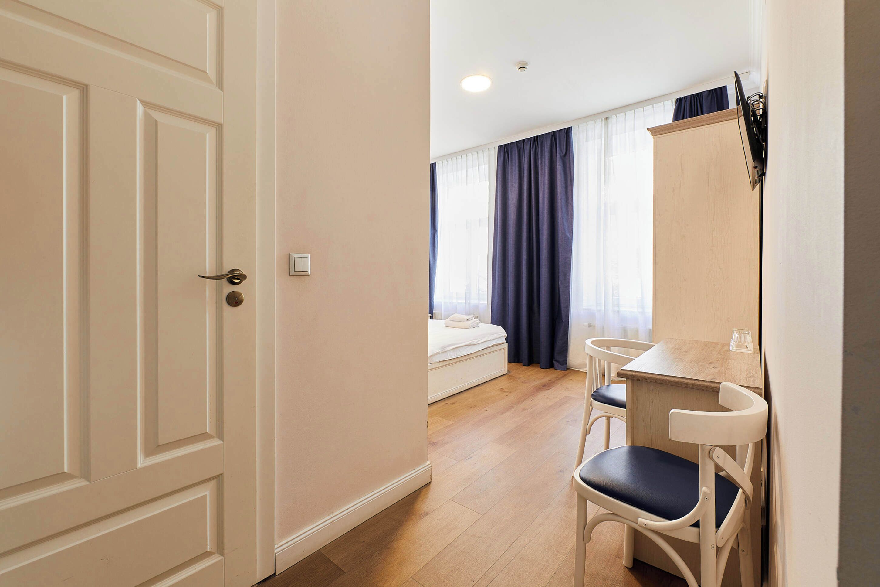 Habitación de hotel con piso de madera, cama con colchón blanco, un escritorio pequeño con silla y un armario.