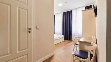 Habitación de hotel con piso de madera, cama con colchón blanco, un escritorio pequeño con silla y un armario.