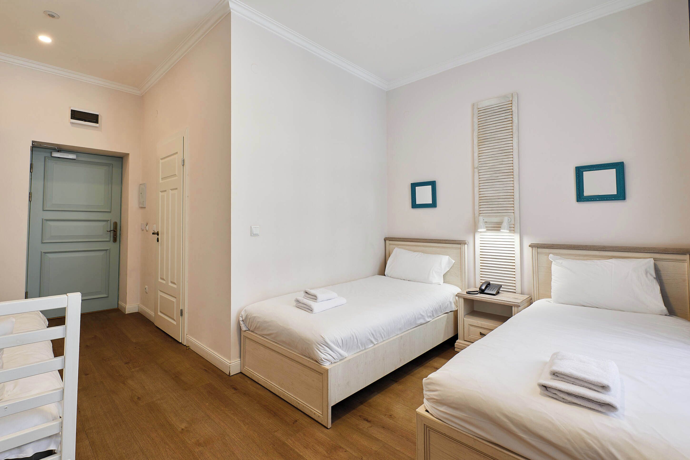 A hotel room with two single beds, a bedside table with a lamp, a small bedside table with a phone, and a door with a light switch.