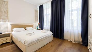 Ein ordentlich bezogenes Bett mit weißen Leinen und Handtüchern, ein Nachttisch mit Lampe und ein Fenster mit dunklen Vorhängen.