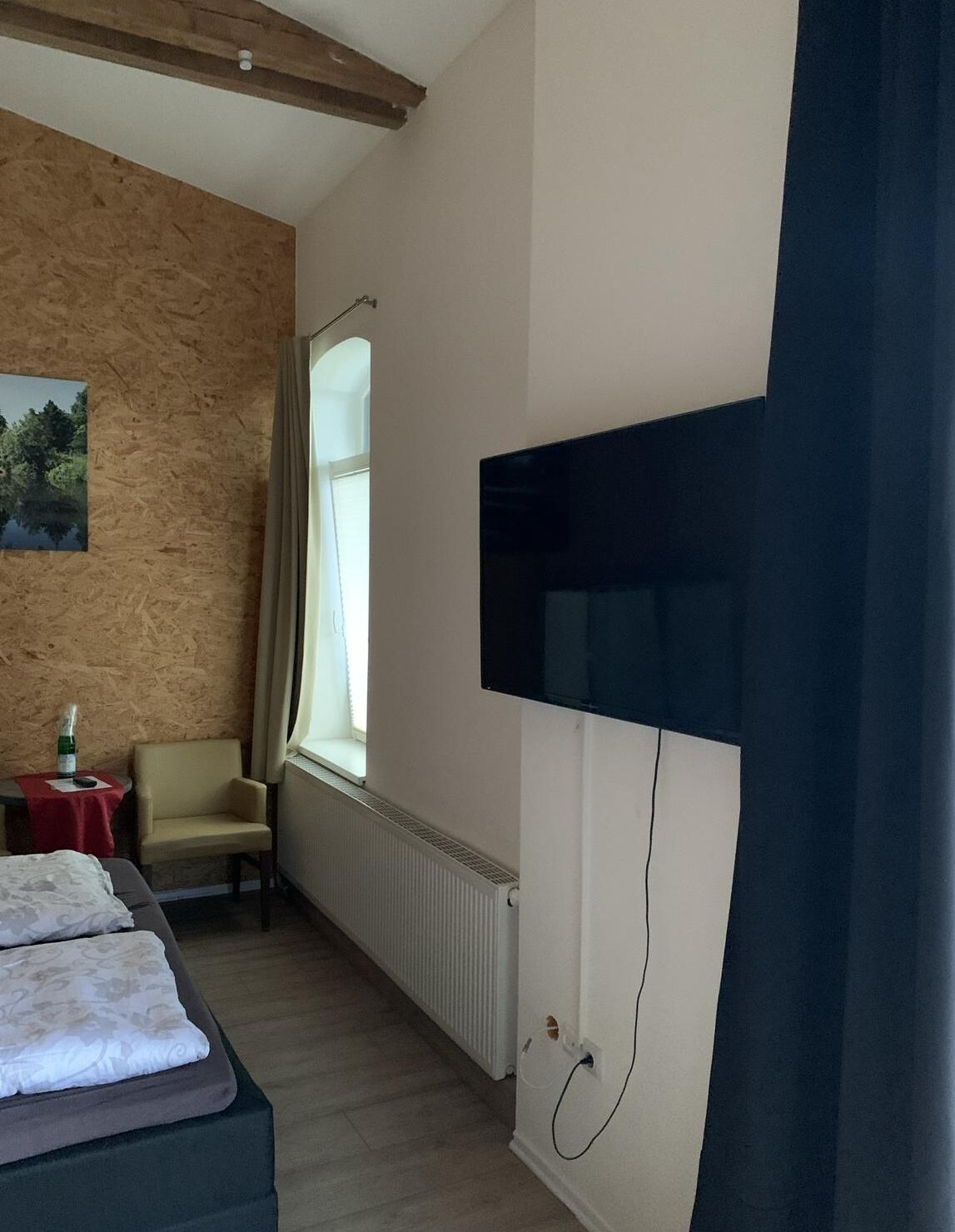 A hotel room with a bed, a chair, a table with a bottle, a window, and a TV mounted on the wall.