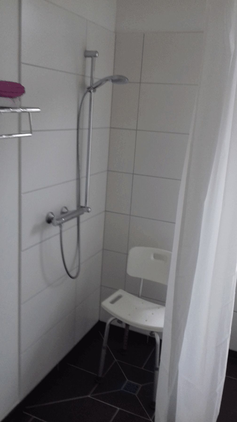 A small bathroom with a shower, a white chair, and a shelf with a pink object.