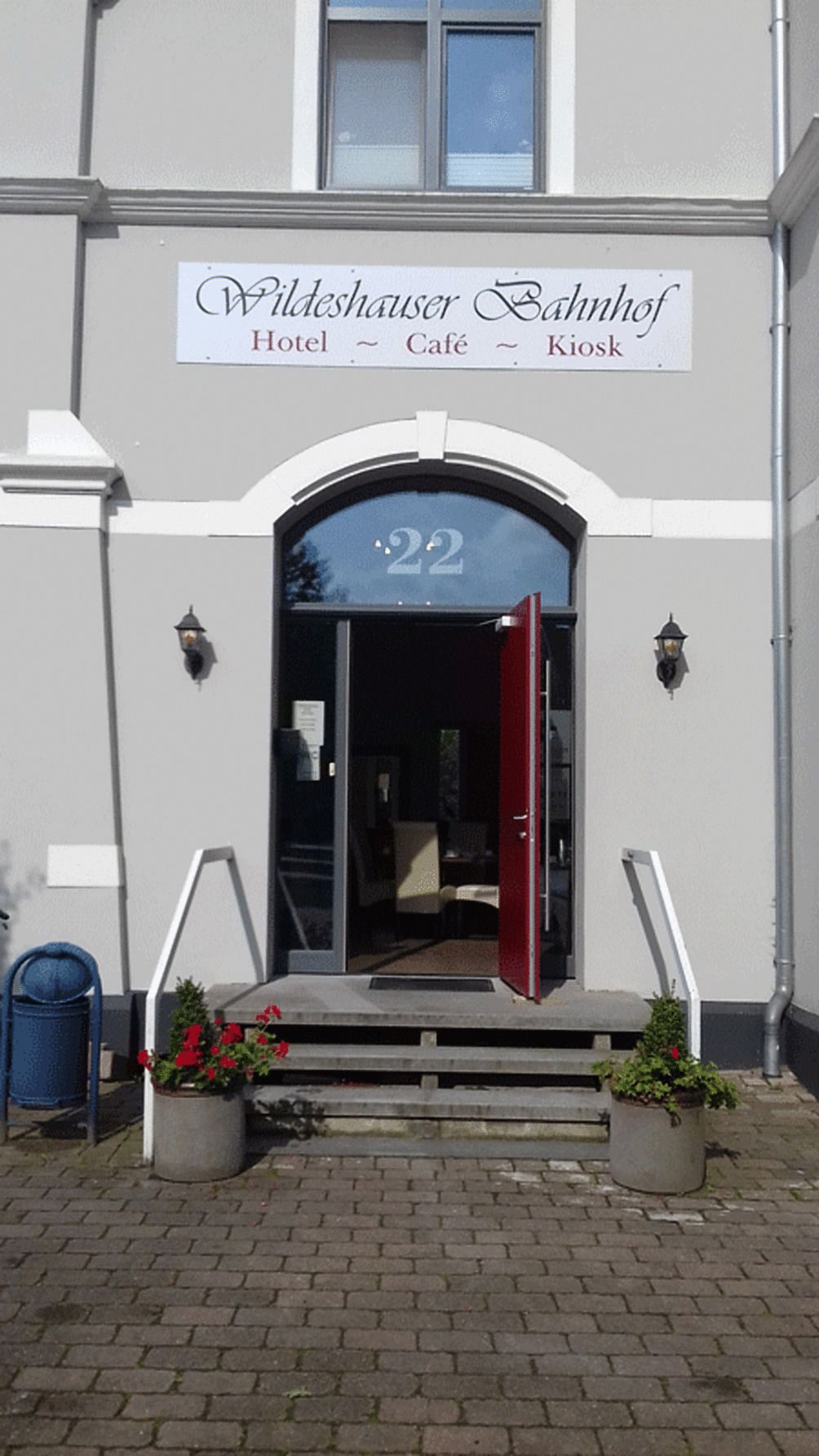 The entrance to the Wildeshaus Bahnhof Hotel, featuring a prominent number '22' above the door, a red door, and two potted plants on the steps.