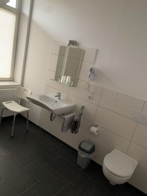 A bathroom with a white sink, mirror, and toilet. There is a window with blinds, a chair, and a trash bin. - Wildeshauser Bahnhof (Wildeshausen)