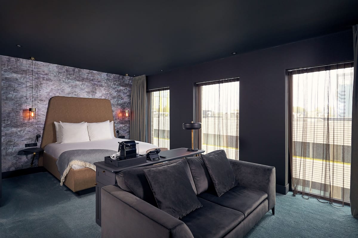 Una habitación de hotel moderna con una cama grande, un sofá y un escritorio. Hay ventanas amplias con cortinas, una lámpara de pared y vistas al exterior.