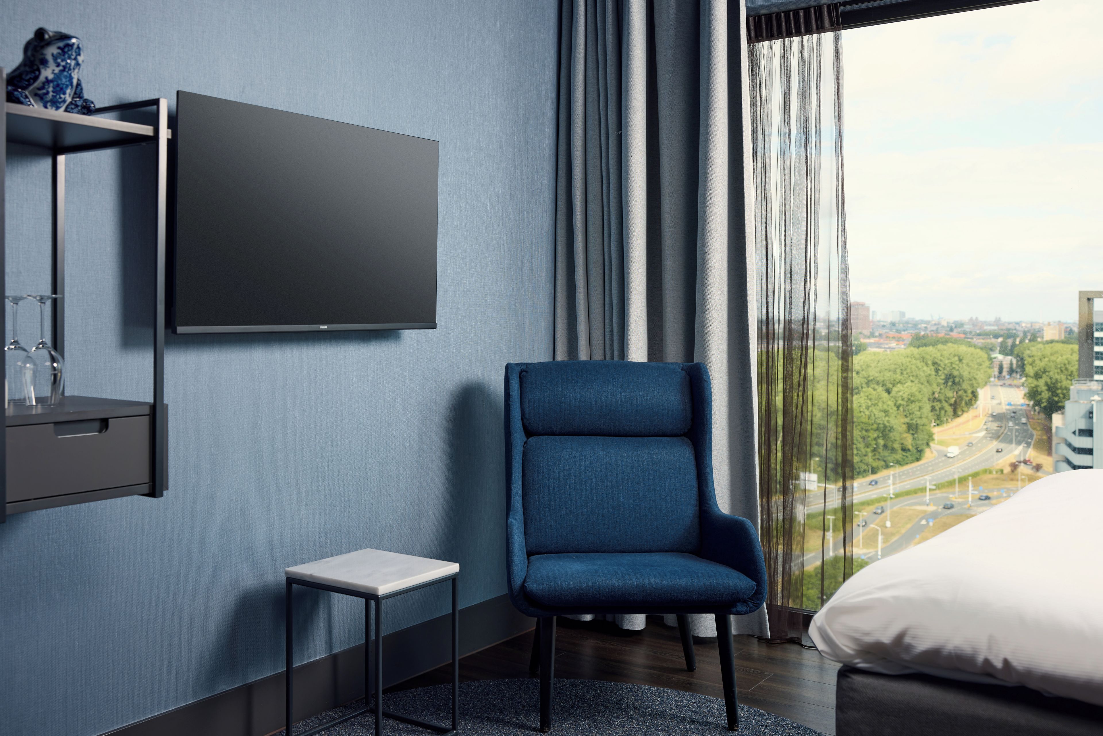 Een moderne hotelkamer met een groot raam, een flatscreen televisie aan de muur, een klein bijzettafeltje, een blauwe fauteuil en een bed met witte beddengoed.