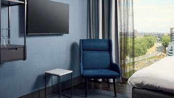 Een moderne hotelkamer met een groot raam, een flatscreen televisie aan de muur, een klein bijzettafeltje, een blauwe fauteuil en een bed met witte beddengoed.