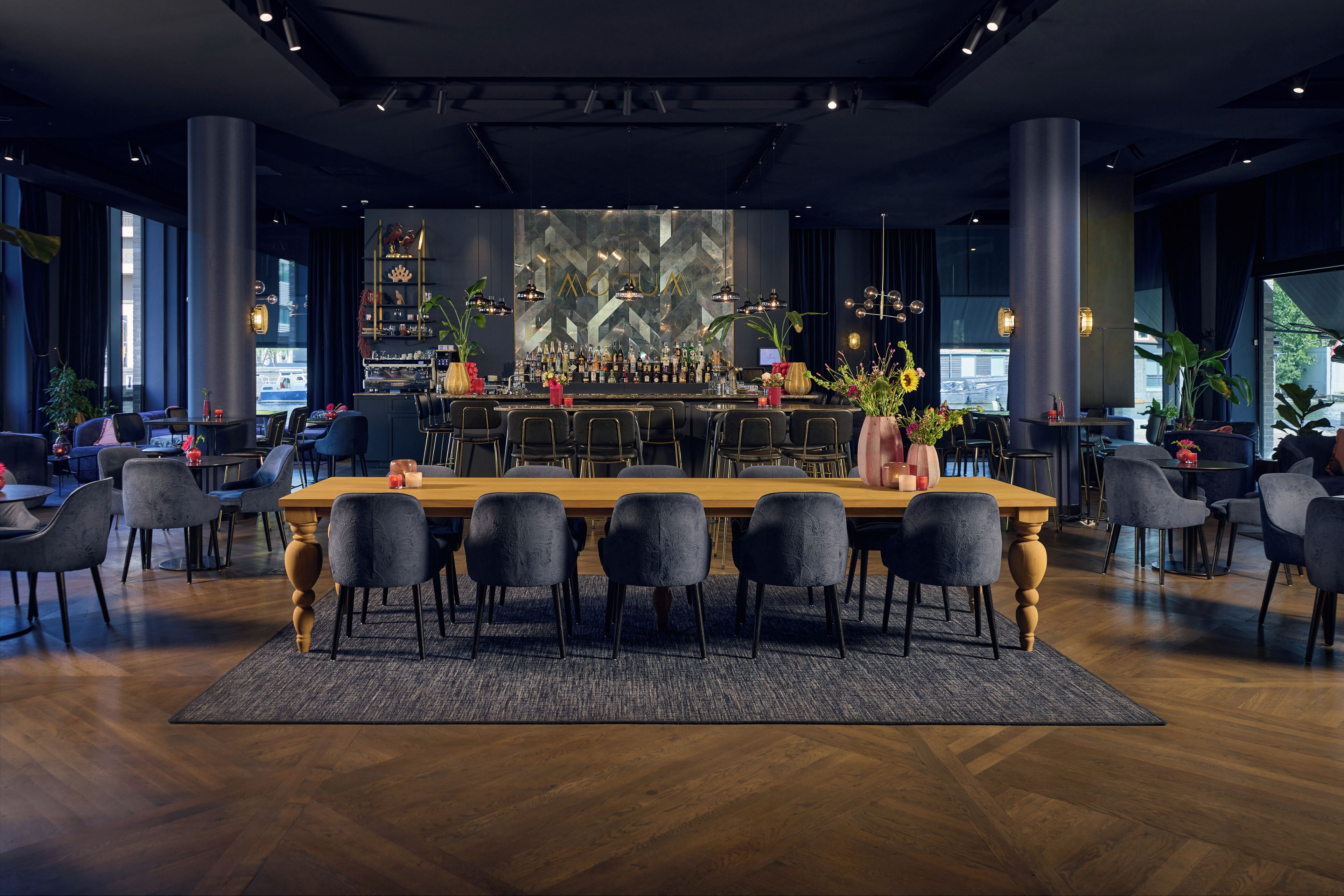 Un área de bar con una mesa de madera larga, varios taburetes altos y una barra repleta de botellas.