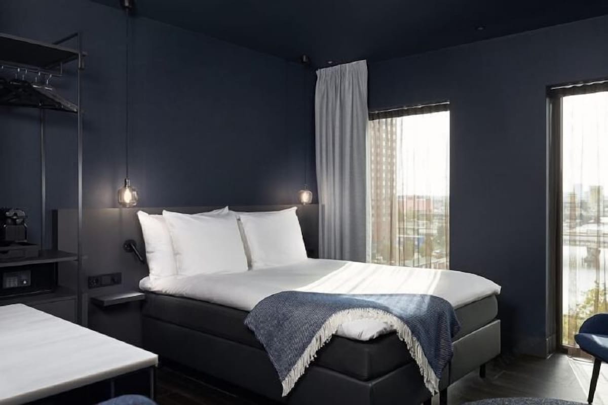 Un dormitorio moderno con una cama, mesitas de noche y vistas de la ciudad a través de grandes ventanales.