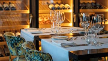 Un comedor con una mesa dispuesta para cuatro personas, rodeada de sillas con tapicería floral. Detrás de la mesa, hay una estantería para vinos con botellas.