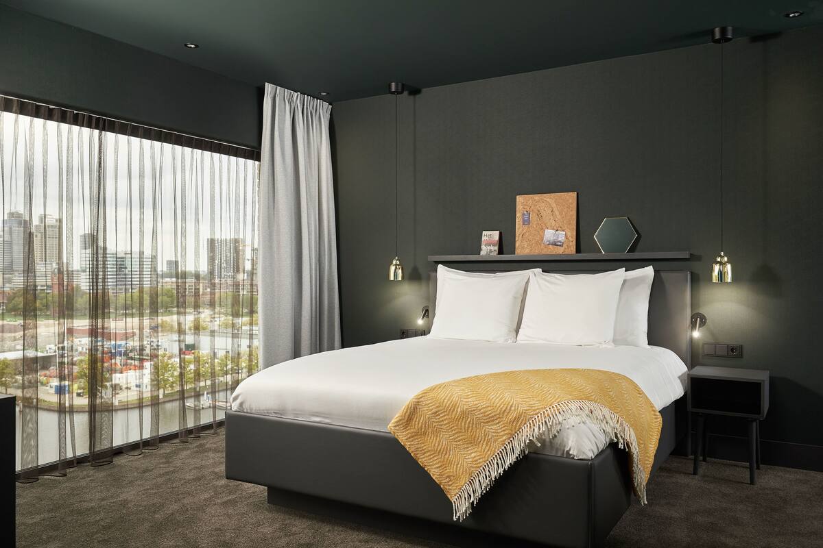 Una habitación de hotel moderna con una cama grande, vista a la ciudad y una decoración en tonos oscuros.